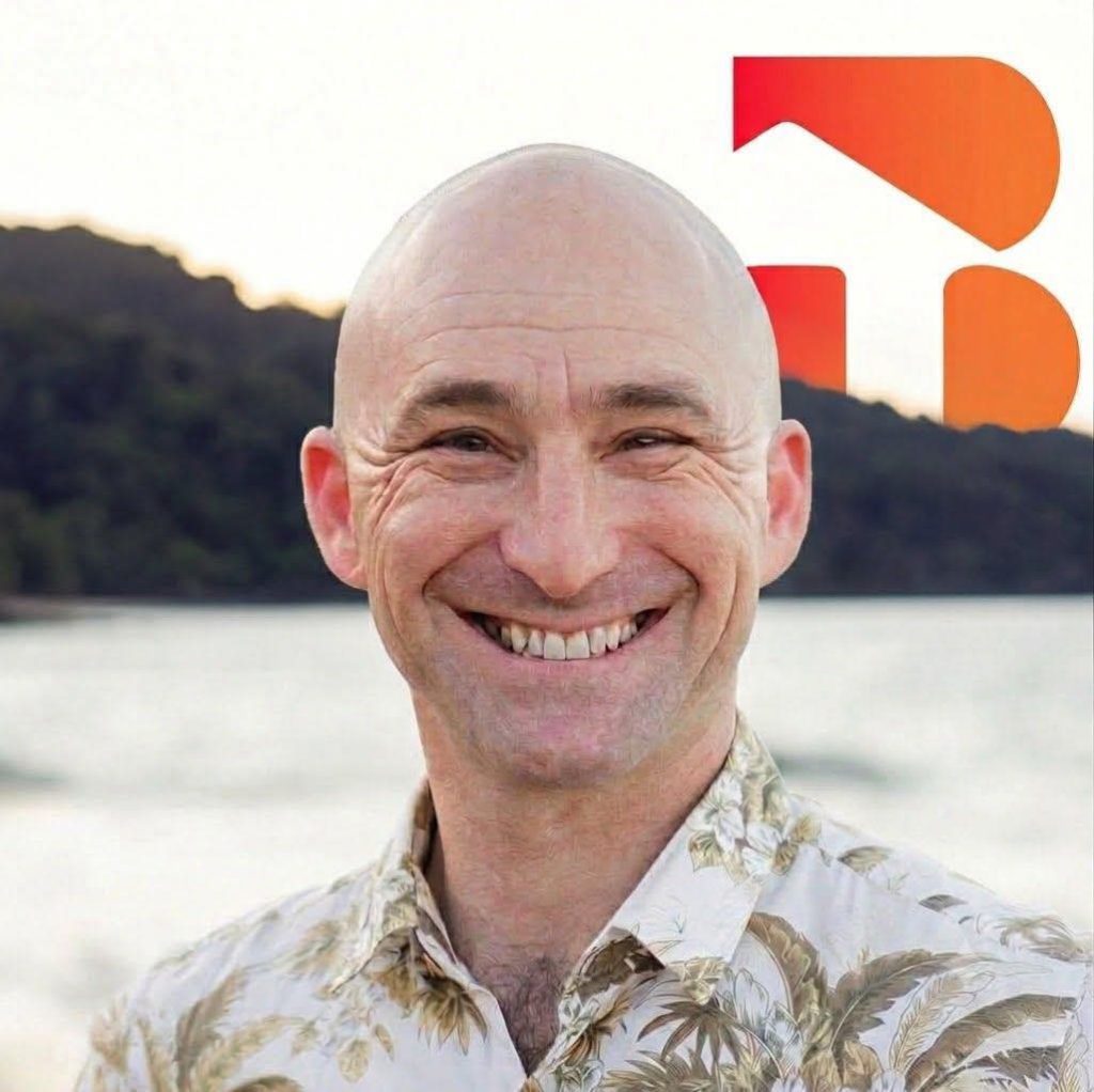 WhatsApp Image 2026-07-22 at 123852 - Bright Property - Bright Property Smiling bald man in a tropical-print shirt at the beach, with water and hills behind and an orange logo in the corner.